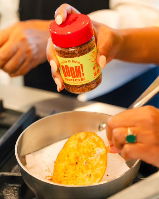 Person holding a jar of 'Boom Spice' over a pan with a tortilla.