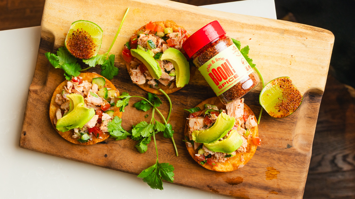 Wooden cutting board with small pizzas topped with avocado and a jar of hot sauce.