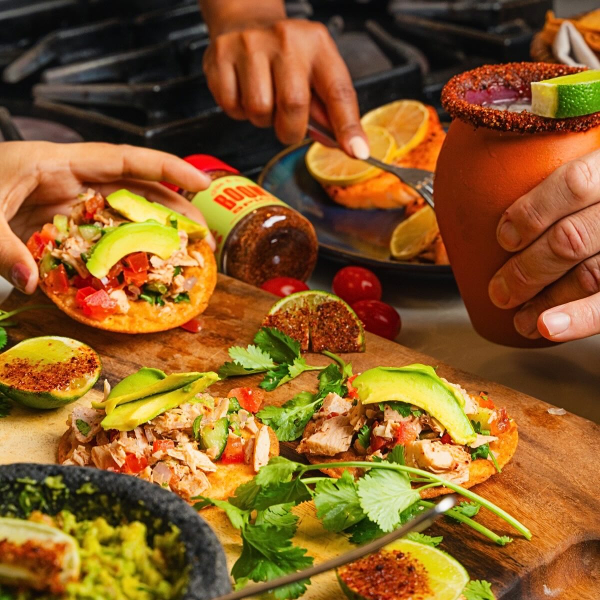 Wooden cutting board with tacos and a jar of hot sauce, surrounded by hands and ingredients.