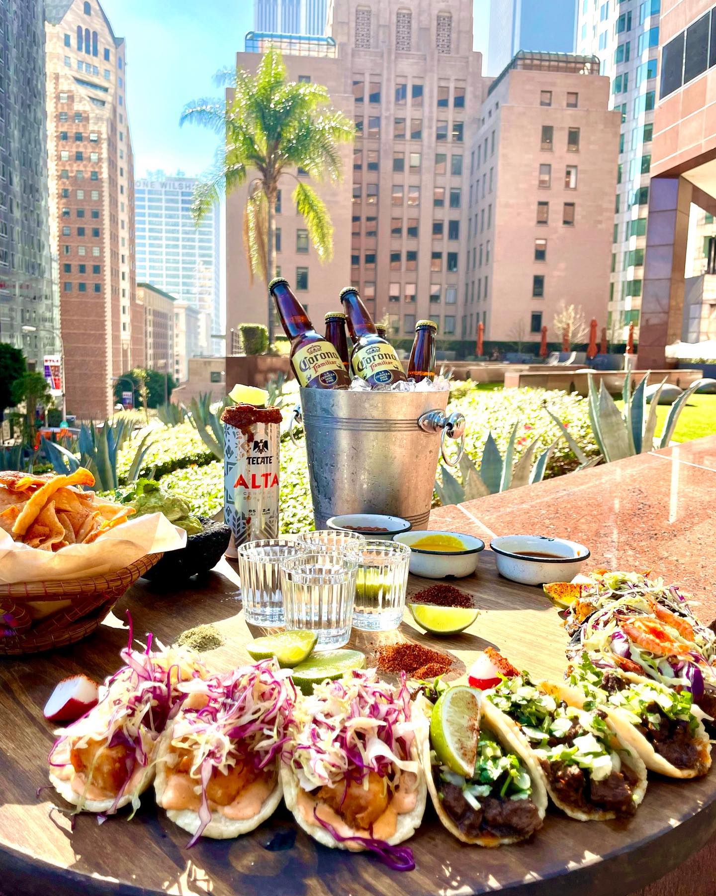 Outdoor dining setup with tacos, drinks, and a cityscape background