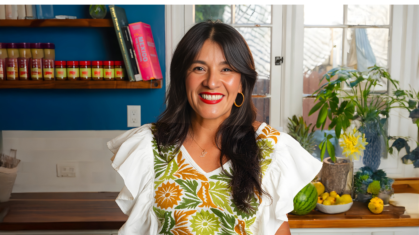 Woman standing in a kitchen with a colorful apron, smiling at the camera.
